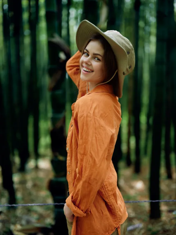 Woman smiling in a hat in a bamboo forest, portrait outdoors wearing an orange linen shirt. Young female model looks over shoulder with joyful grin and relaxed natural lifestyle vibe.