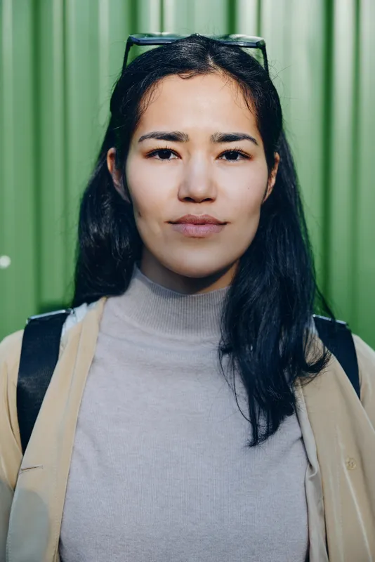 Woman with glasses, casual attire, backpack, stands before a green wall, confident expression, relaxed posture, urban exploration style, everyday travel vibe, approachable and ready