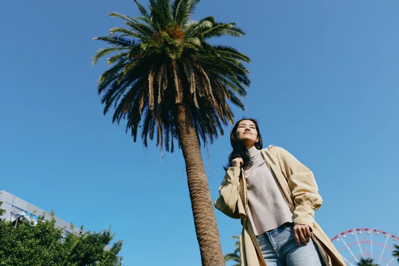 Woman standing beneath a tall palm tree in a sunny outdoor park, casual fashion and relaxed pose, urban greenery and blue sky creating a vibrant lifestyle scene