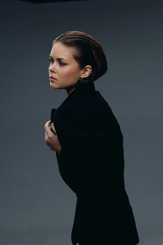 Young woman in black coat looking thoughtful against dark background, moody and stylish portrait.