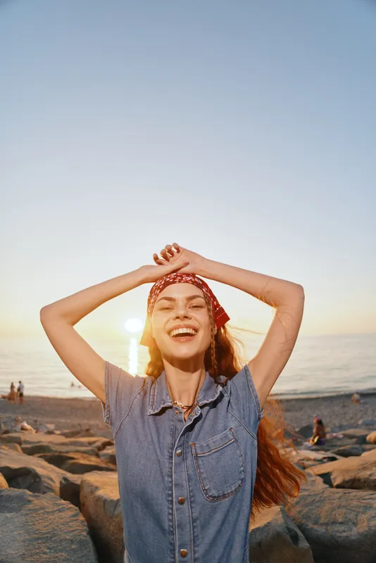 Smiling woman enjoying sunset at rocky beach wearing casual denim shirt and red headscarf with golden light and clear sky in warm, joyful atmosphere.