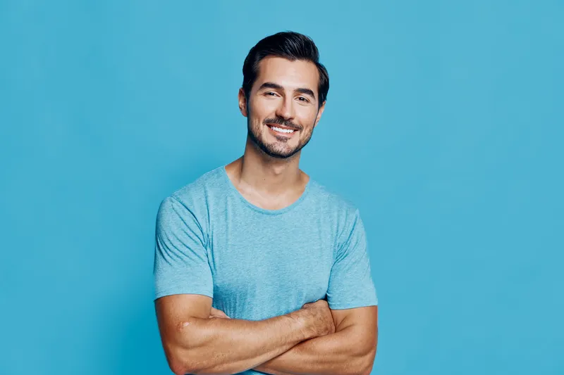 Smiling man with crossed arms wearing a casual blue t-shirt stands against a solid turquoise background, exuding confidence and approachability. People lifestyle concept