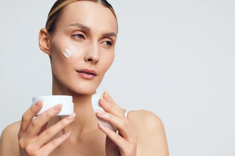 Woman applying moisturizer skincare cream on her face, holding a small jar, calm beauty routine, soft studio light, smooth skin with hydration, white background, close up, serene expression