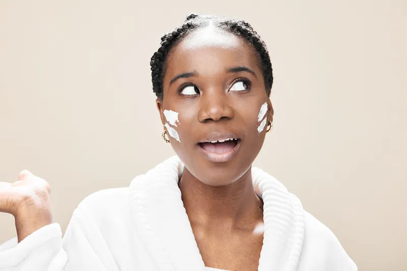 Cheerful Black woman applying skincare cream, showcasing a glowing complexion, casual white bathrobe, and a light, neutral background for wellness and beauty themes