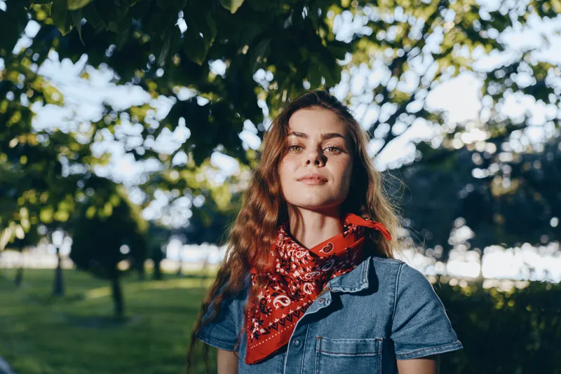 Young woman with red bandana and denim shirt, standing outdoors in a sunlit park with green trees and bright natural light, enjoying a peaceful moment.