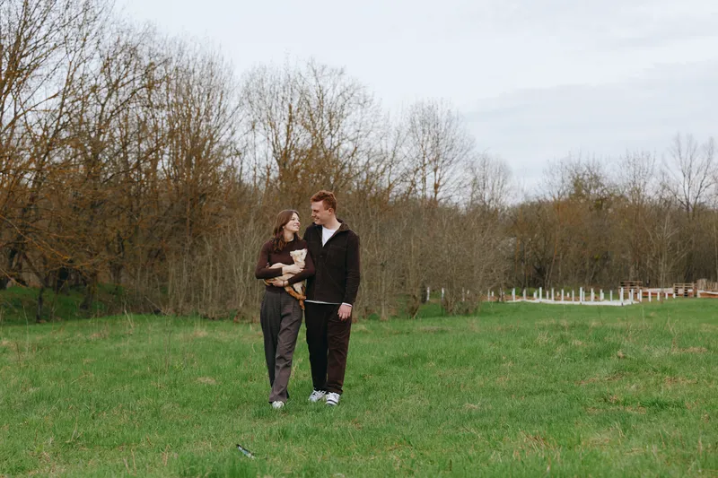 A couple in a grassy field with a small goat kid, sharing a quiet moment outdoors. A warm lifestyle scene highlighting family bonds, rural serenity, and gentle animal friendship.