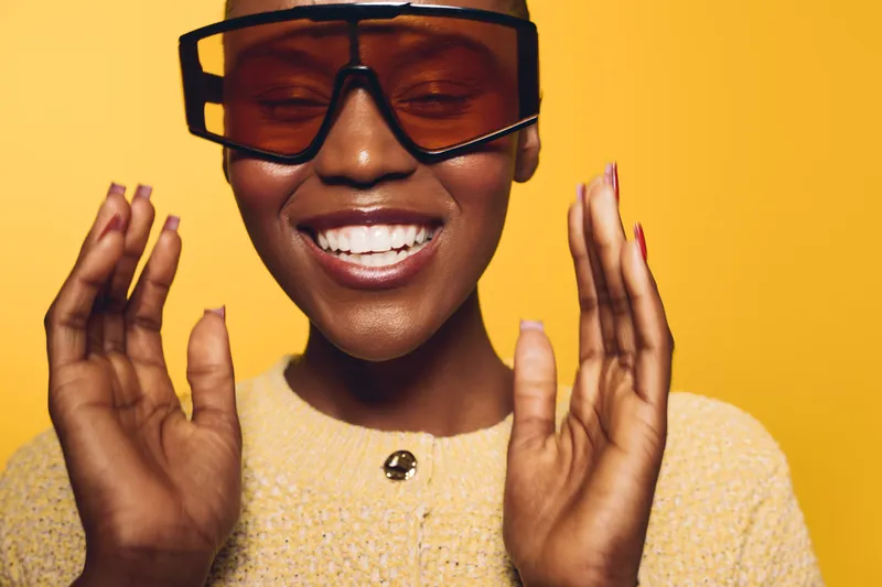 Smiling African woman with dark skin wearing large glasses and a yellow sweater, expressing joy with closed eyes and raised hands. Studio portrait on yellow background.