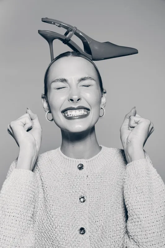 Smiling white woman with light skin closes eyes and clenches fists happily with a high heel shoe balanced on her head. Concept for joy, fashion, and playful mood.