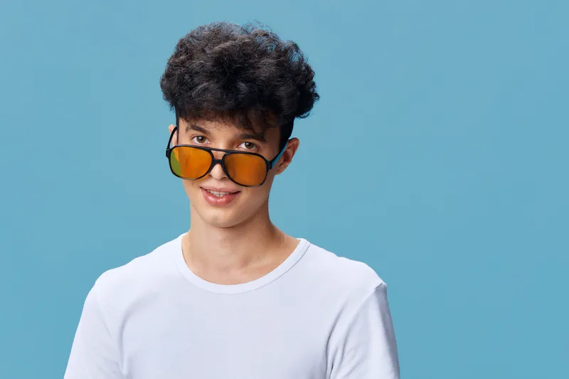 young man wearing orange sunglasses and white t-shirt smiling against blue background with curly hair and casual style, vibrant and modern portrait