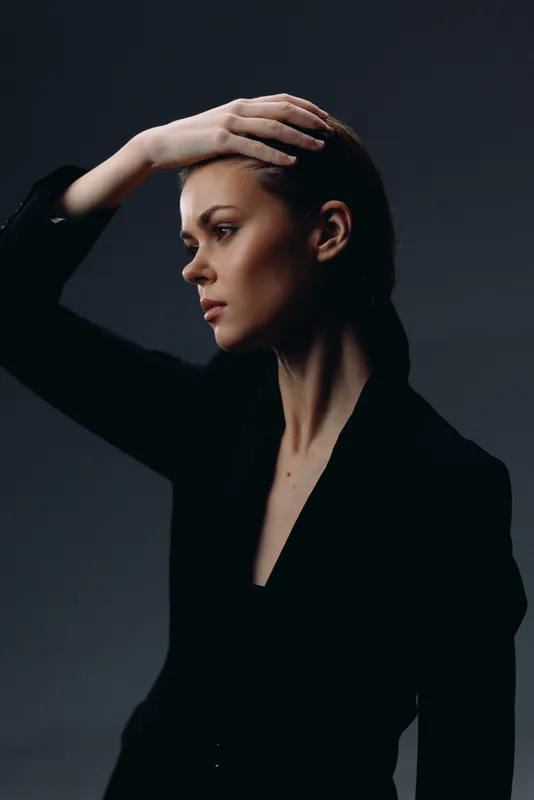 Thoughtful woman in black outfit with hand on head against dark background, moody portrait, focused and elegant expression
