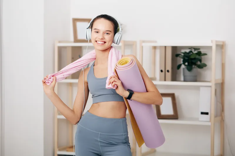 Active woman fitness enthusiast with headphones, carrying a yoga mat and towel in a bright home gym, ready for a workout, energetic, focused and stylish in athleisure.