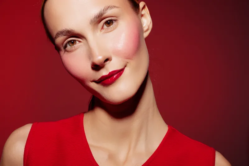 Close-up of a smiling woman with red lipstick and blush wearing a red sleeveless top against a red background. Concept of beauty, makeup, fashion, and confidence.