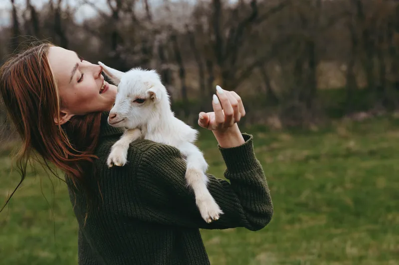 Young woman with red hair happily holds a white baby goat on her shoulder outdoors in a natural green setting during a casual lifestyle moment.