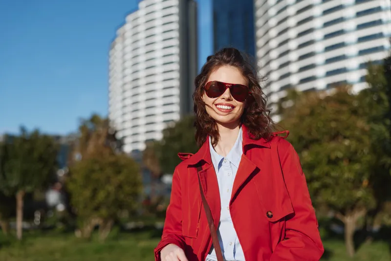 One woman with brunette hair wears a redcoat and sunglasses, smiling in a sunny park with a modern cityscape in the background.