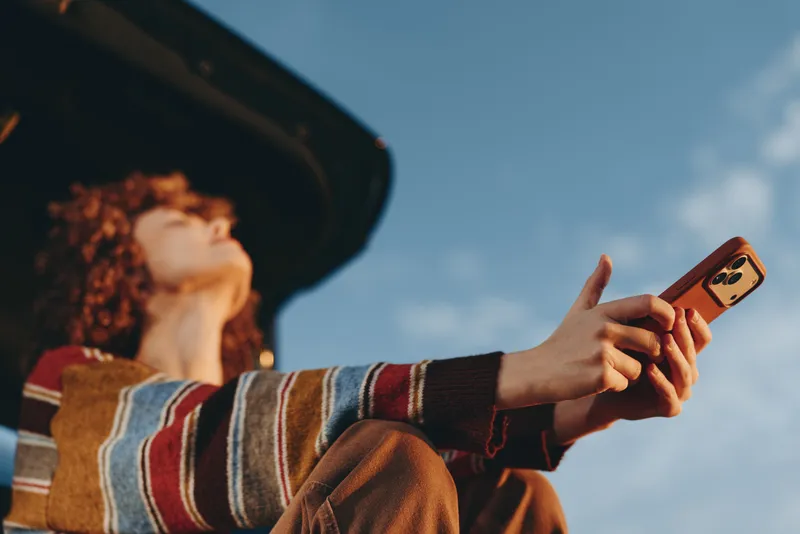 Woman hands hold smartphone during road travel lifestyle outdoors in daylight, casual striped sweater, passenger seat view, peaceful communication moment, sky sunlit background