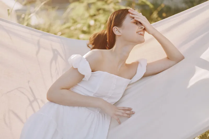 Young woman in a delicate white dress enjoying a sunny day outdoors, expressing joy and relaxation in a serene natural setting, surrounded by soft greenery