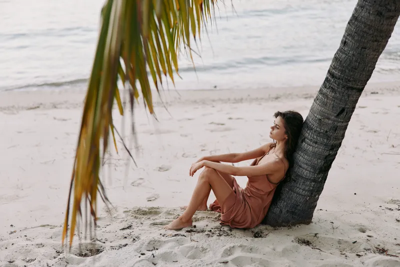 A woman sits against a leaning palm on a sandy beach, eyes closed, enjoying warm light and a gentle breeze as calm waves kiss the shore and distant horizon glows softly.
