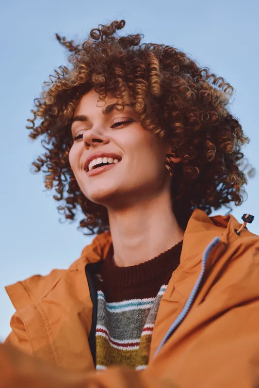 Young woman lifestyle portrait, curly hair, orange jacket, smiling outdoors under clear blue sky, natural light on face, relaxed casual mood for happiness and freedom concept