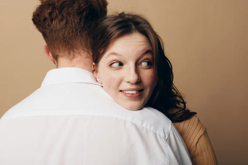 Young woman with freckles smiles softly while hugging a man in white shirt, looking sideways. Intimate moment, affection, and connection between couple in warm studio setting.