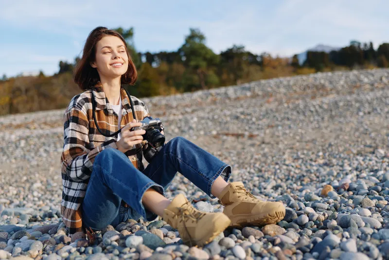 Woman photographer sitting on a pebble beach with a camera, wearing a plaid shirt, jeans and boots, enjoying outdoor scenery and a creative moment by the shore
