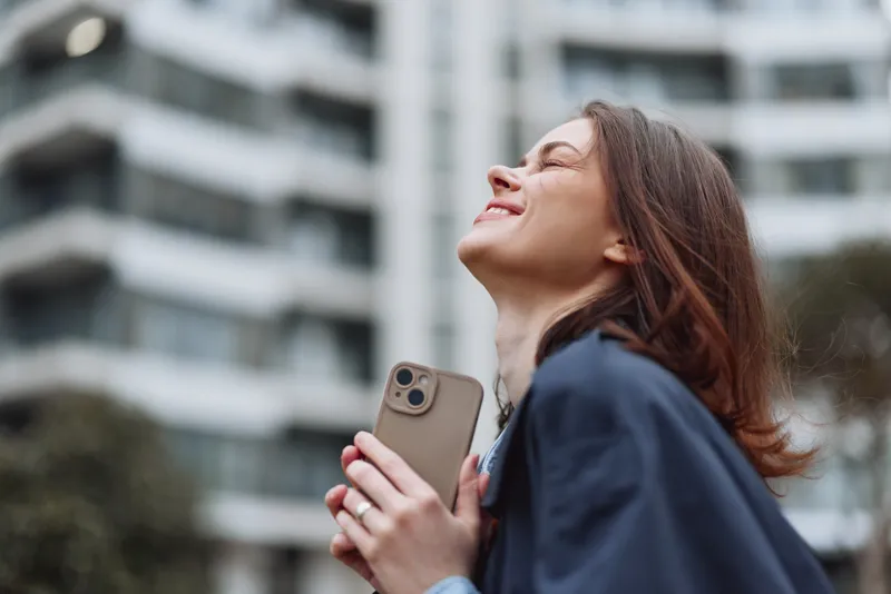 A smiling woman with a smartphone stands outdoors in an urban setting, basking in daylight as glass buildings shimmer behind her, suggesting urban connectivity.