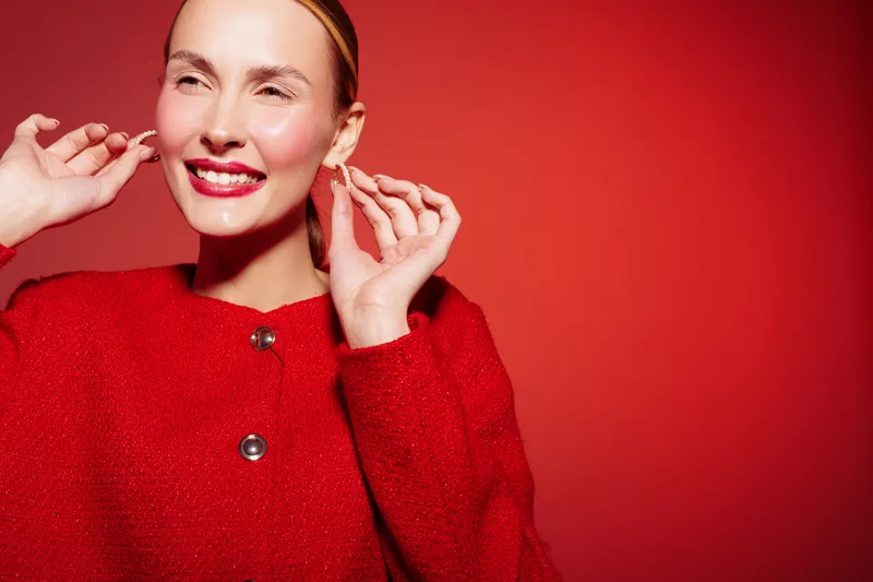 Smiling woman in red coat adjusts earrings against red background. Bright makeup and joyful expression create festive and elegant mood. Concept of fashion, beauty, and celebration.
