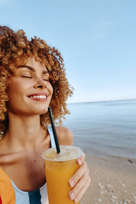Cheerful woman on a sunny beach enjoying a bright juice drink, eyes closed, relaxed posture, sea breeze, warm light, natural colors, coastal background