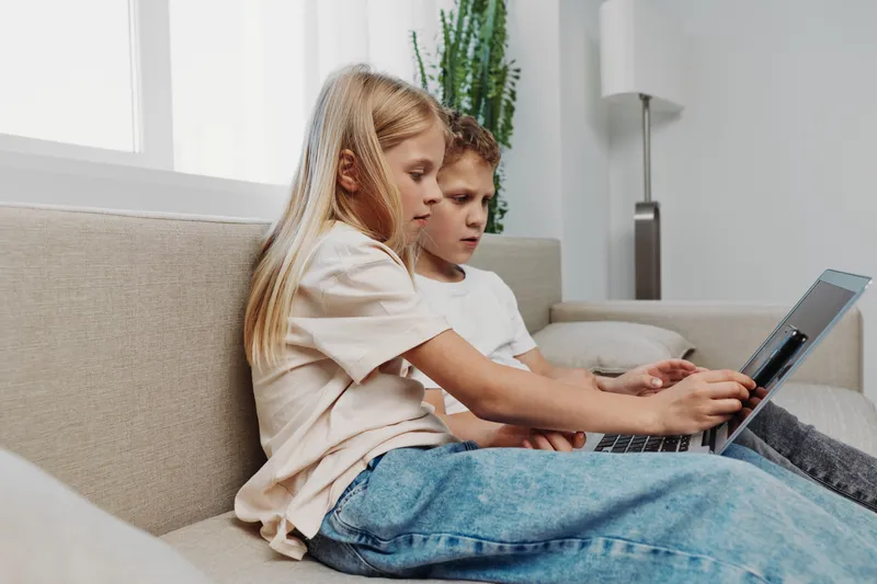 Two focused children sit on a beige sofa using a laptop together in a bright modern living room. Concept of learning, technology, cooperation, and childhood education at home.
