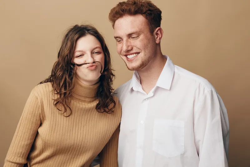 Young couple having fun together, woman making a mustache with her hair while man smiles. Casual clothing and warm beige background create a relaxed and playful mood.