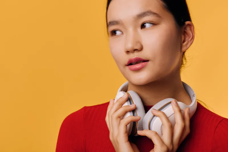 Woman with headphones and red sweater looking aside on orange background. Portrait of thoughtful female with earphones for music or relaxation in bright studio light.