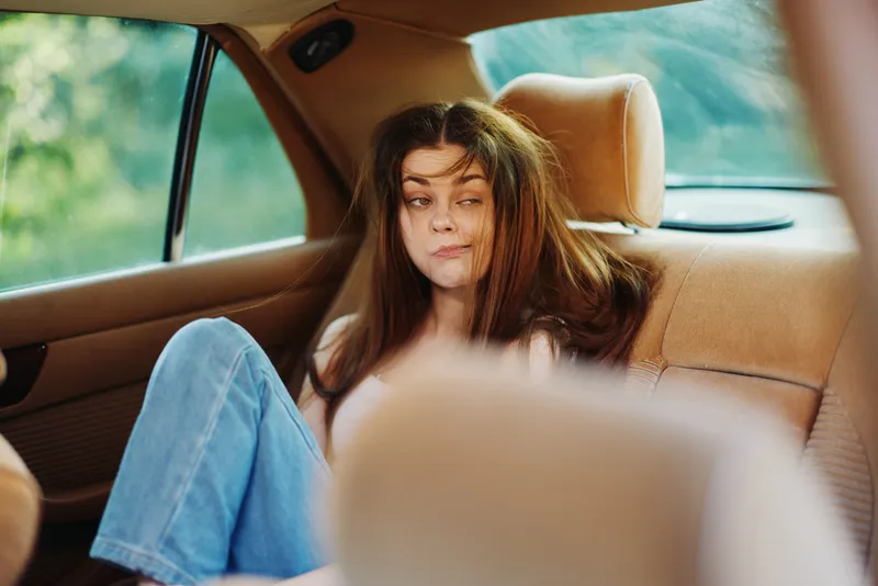 A young woman with messy hair sitting in a car, expressing boredom or frustration in a casual outfit, capturing a relatable moment of travel discomfort.