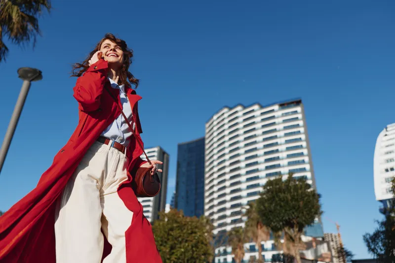 Young woman in a red coat walks confidently through a sunny urban street, smiling as tall modern buildings rise behind her, creating a vibrant city fashion moment.
