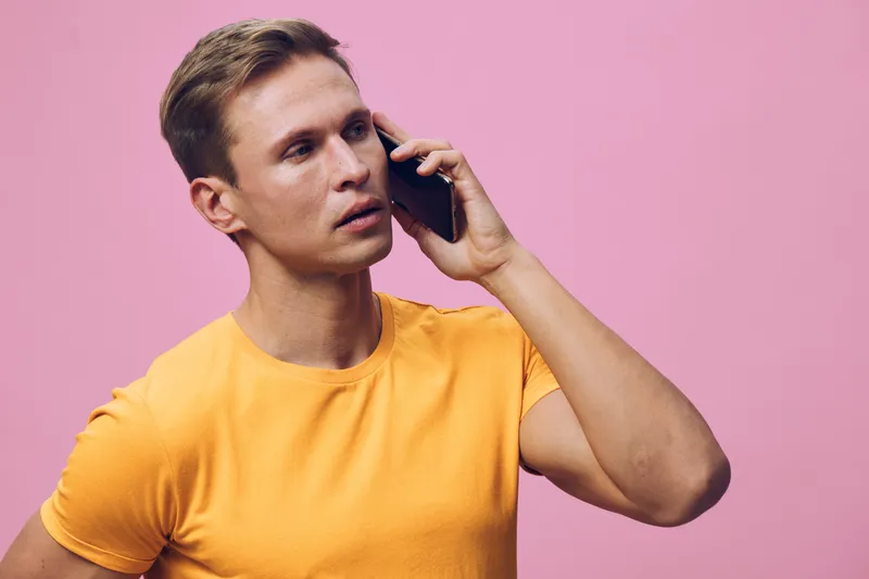 young man talking smartphone serious facial expression wear orange t-shirt isolated pink background communication digital nomad concept