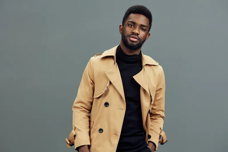 Stylish young Black man wearing a beige trench coat and black turtleneck against a muted gray background, exuding confidence and sophistication