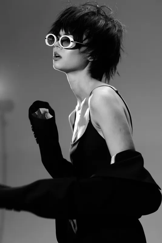 Black and white woman wearing stylish sunglasses and dark clothing posing with confident expression in studio lighting.