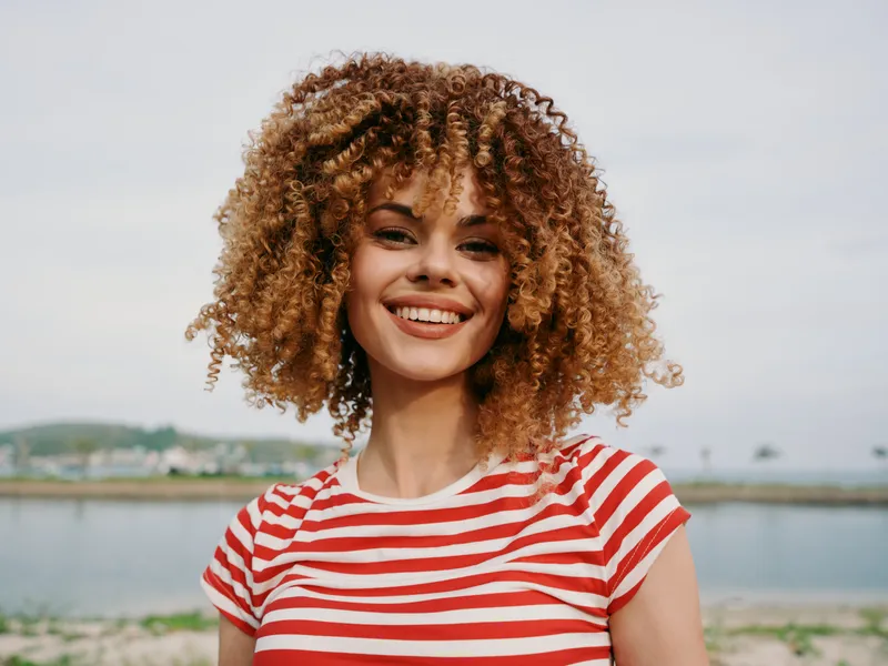 Closeup portrait of a smiling young woman with curly hair wearing a red striped shirt, outdoors near calm water, capturing bright confidence, natural charm, and warm sunlight.