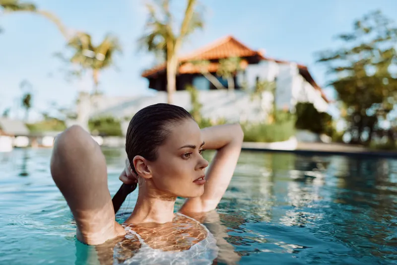 Woman relaxing in a pool, sunlit water, outdoor resort setting, lifestyle focus on leisure and self care, calm refreshing mood, natural light and relaxed vibe.