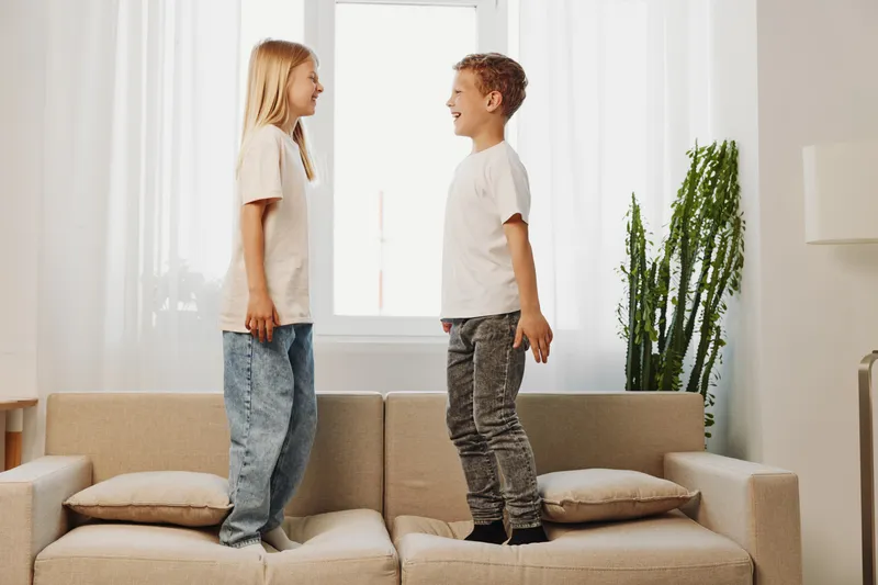 Two happy children stand facing each other on a beige sofa in a bright living room with casual clothes. Concept of childhood joy, friendship, and playful indoor moments.
