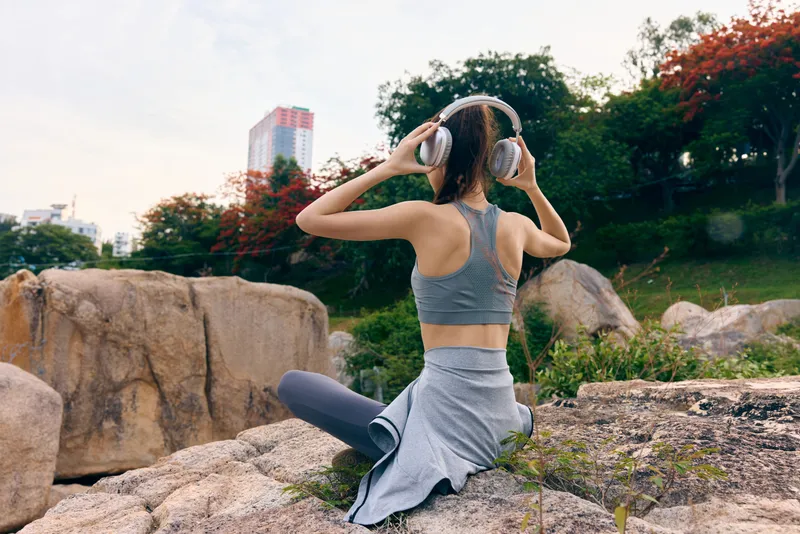 Young woman in sportswear enjoying music outdoors on a rock during sunset, emphasizing wellness and fitness with a vibrant natural background