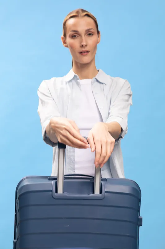 Young woman in casual clothes holding a blue suitcase handle with both hands. Studio portrait on blue background. Concept of travel, journey, and vacation preparation.