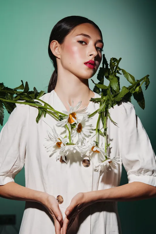 Fashionable woman with glowy skin and bold red lips wears white couture adorned with wild daisies on green stems. Studio portrait with artistic lighting emphasizes beauty and creative concept.