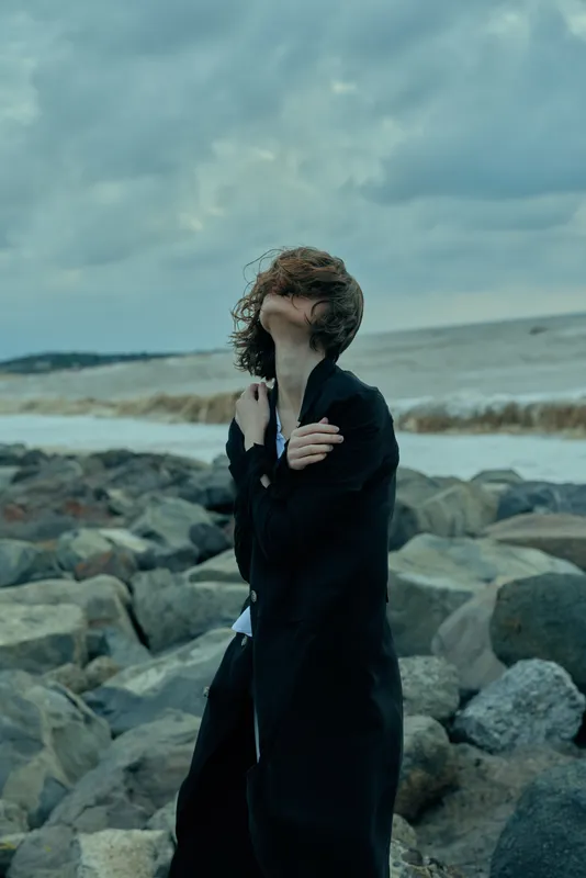 Windy seascape with a cloaked figure on rocky shore, hair blown across face, moody atmosphere, dark coat, ocean backdrop, rough water, dramatic weather, solitary stance, artful silhouette, emotional