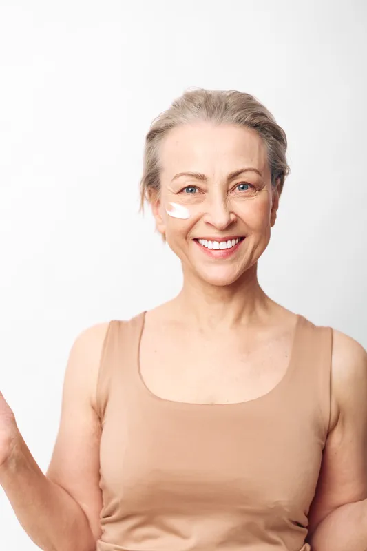 Smiling mature woman with short hair applies white facial cream on cheek, wearing beige tank top, isolated on white background. Concept of skincare, beauty, self-care, and healthy aging.