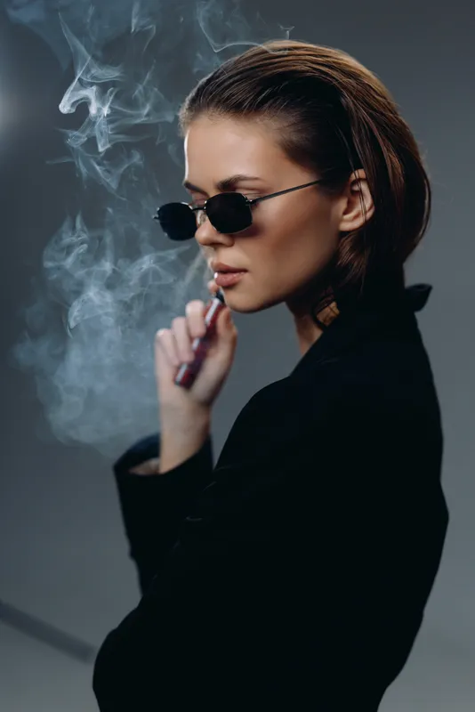 Stylish woman smoking vape wearing sunglasses and dark clothing with smoke around her in low light atmosphere