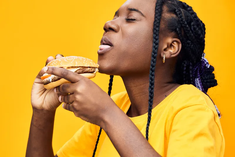 Joyful young woman enjoying a delicious burger against a vibrant yellow background, showcasing a moment of happiness and satisfaction