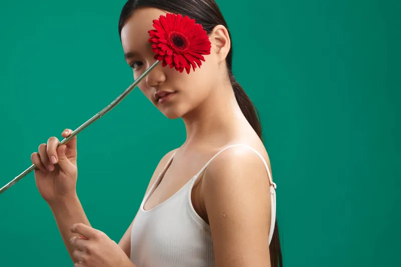 asian woman poses with a red flower held beside her eye against a vivid green backdrop, creating a bold artistic portrait that exudes calm elegance and modern studio aesthetics.
