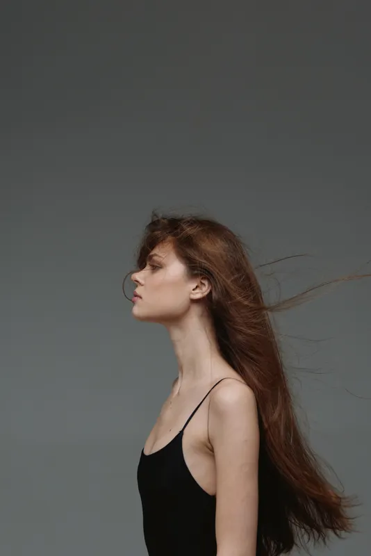 Profile of a slender woman with long red hair wearing a black top against a gray background, showing calm and serene expression.