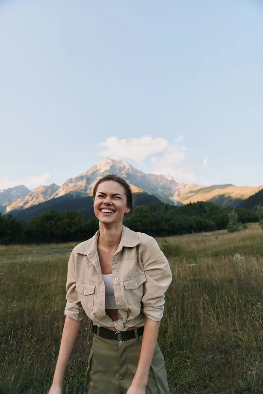 Young woman stands in a grassy field with distant mountains, wearing a beige jacket over a white top and olive pants, smiling softly in bright outdoor light.