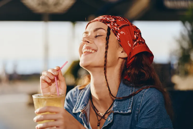 lifestyle woman in modern boho-western denim street style enjoys refreshing drink with joyful expression under warm sunlight, film color effect enhancing natural tones