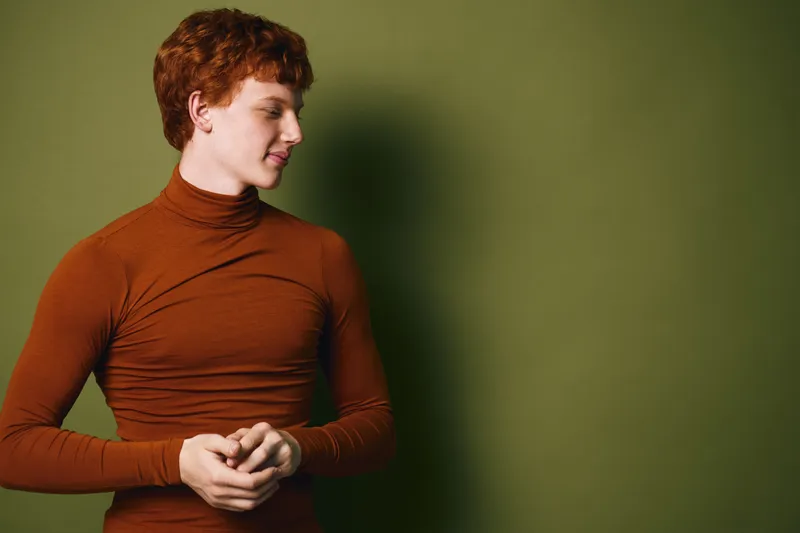 Young woman with red hair in rust turtleneck sweater looks aside thoughtfully against green background. Portrait with natural light and casual confident pose in simple indoor setting.